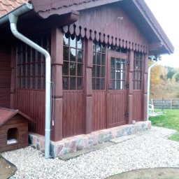 TOMASZEWSKI MEBLE PAWEŁ TOMASZEWSKI - Drewniana weranda z oknami i drzwiami, ozdobne wykończenie dachu, brązowa kolorystyka, kamienna podmurówka i żwirowe podłoże. Obok buda dla psa.