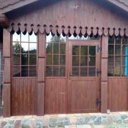 TOMASZEWSKI MEBLE PAWEŁ TOMASZEWSKI - Drewniana altana z oknami i drzwiami w rustykalnym stylu, z dekoracyjnym wykończeniem dachu i kamienną podmurówką. Widoczne detale stolarskie i naturalne otoczenie.