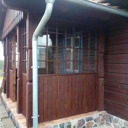 TOMASZEWSKI MEBLE PAWEŁ TOMASZEWSKI - Zadaszona weranda z drewnianą konstrukcją, brązowymi panelami i oknami z niebieskimi ramami. Widoczny fragment rynny i kamienny fundament.