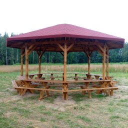 TOMASZEWSKI MEBLE PAWEŁ TOMASZEWSKI - Drewniana altana z czerwonym dachem, otoczona stołami i ławkami, na tle łąki i lasu. Widoczne detale konstrukcyjne i naturalne otoczenie.