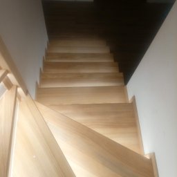 TOMASZEWSKI MEBLE PAWEŁ TOMASZEWSKI - Jasne, drewniane schody z widoczną strukturą drewna, prowadzące w górę, z białymi ścianami po bokach i ciemnym sufitem w tle. Ujęcie z góry, podkreślające naturalny materiał.
