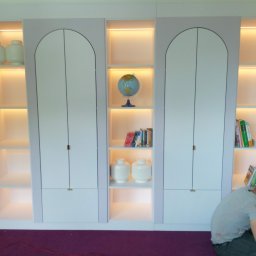 TOMASZEWSKI MEBLE PAWEŁ TOMASZEWSKI - Biała szafa wnękowa z oświetleniem LED i zaokrąglonymi drzwiami, uzupełniona dekoracjami. Montażysta w trakcie pracy, widoczny na pierwszym planie.