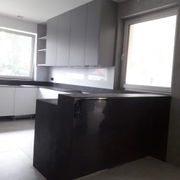 TOMASZEWSKI MEBLE PAWEŁ TOMASZEWSKI - Nowoczesna kuchnia z szarymi szafkami i czarnym blatem, widok na wyspę kuchenną. Minimalistyczny design, jasne wnętrze z oknami.