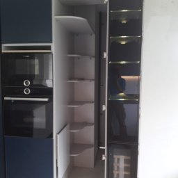 TOMASZEWSKI MEBLE PAWEŁ TOMASZEWSKI - Nowoczesna zabudowa kuchenna z piekarnikiem, szafkami w kolorze granatowym i białym, oraz otwartą szafką z półkami i szafką na wino z czarnymi, szklanymi drzwiami.