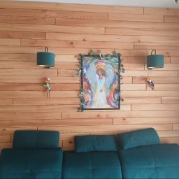 Drewniana ściana z poziomych desek, ozdobiona obrazem w ramie i lampkami z motywem papug. Pod ścianą zielona sofa.