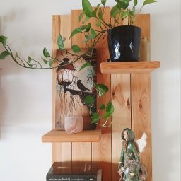 Drewniana półka z desek z trzema poziomami, na których stoją książki, roślina doniczkowa, figurka anioła i obrazek z motywem latarni i kruka. Naturalne drewno, jasne tło.