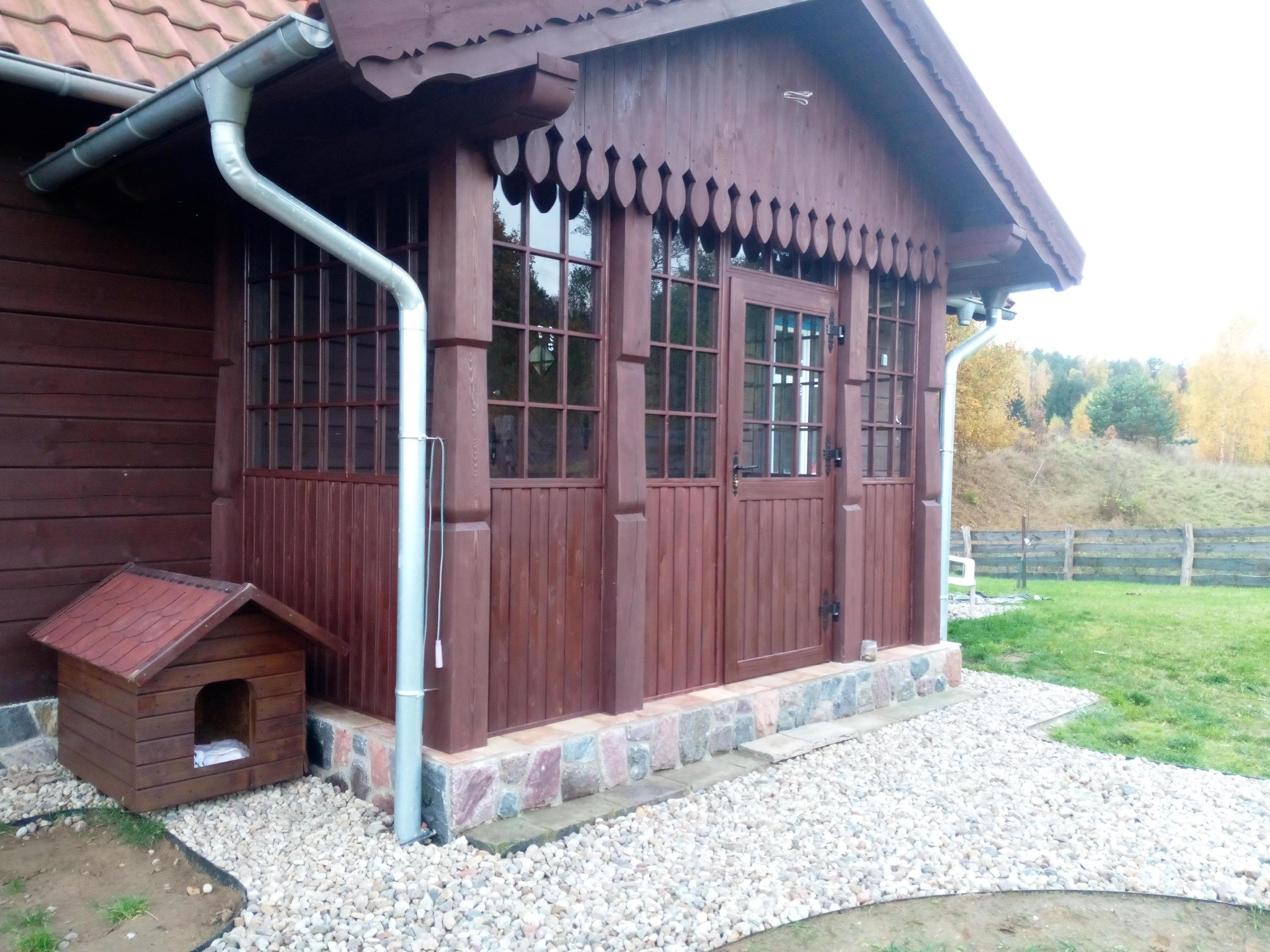 Drewniana weranda z oknami i drzwiami, ozdobne wykończenie dachu, brązowa kolorystyka, kamienna podmurówka i żwirowe podłoże. Obok buda dla psa.