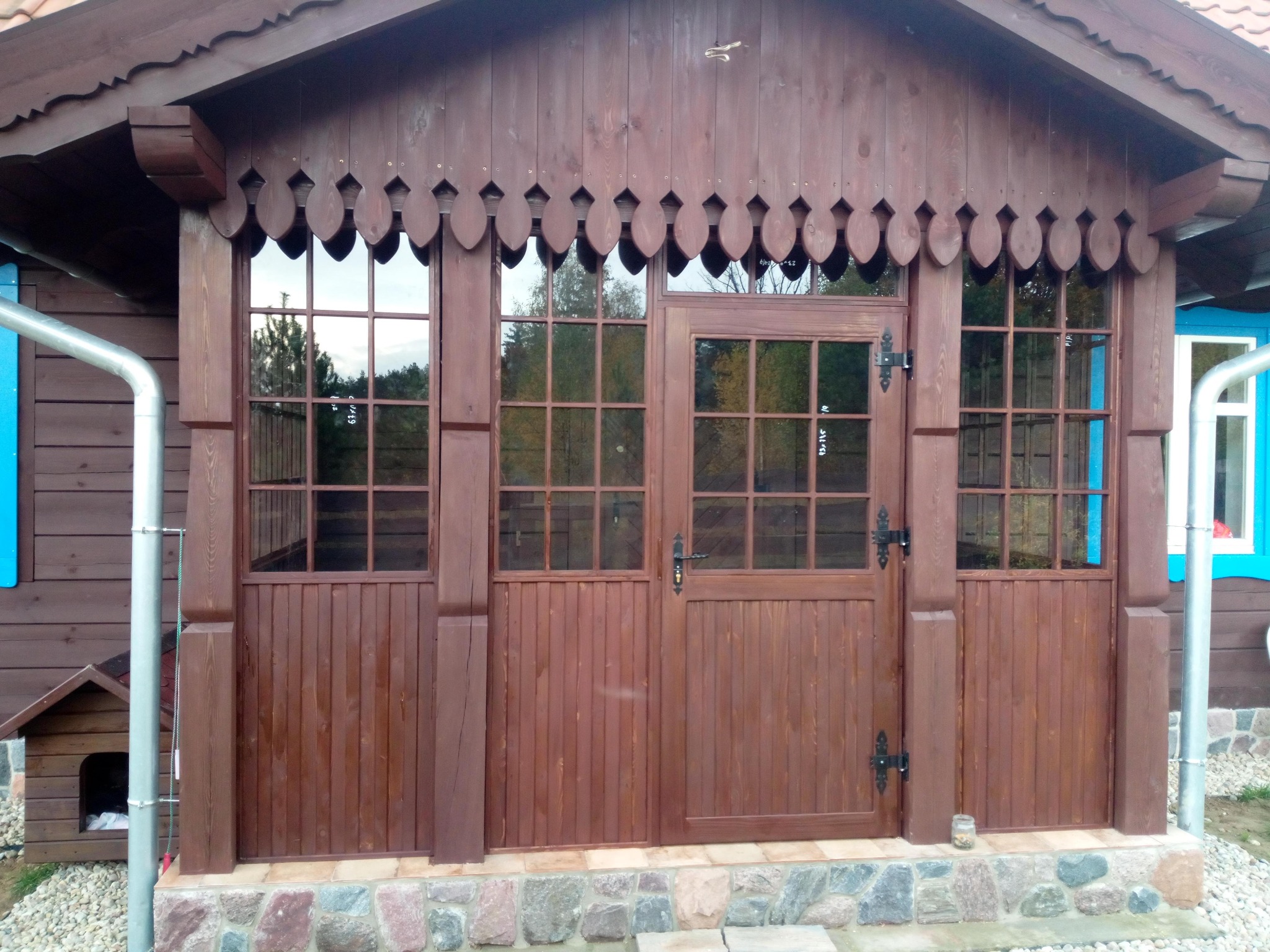 Drewniana altana z oknami i drzwiami w rustykalnym stylu, z dekoracyjnym wykończeniem dachu i kamienną podmurówką. Widoczne detale stolarskie i naturalne otoczenie.