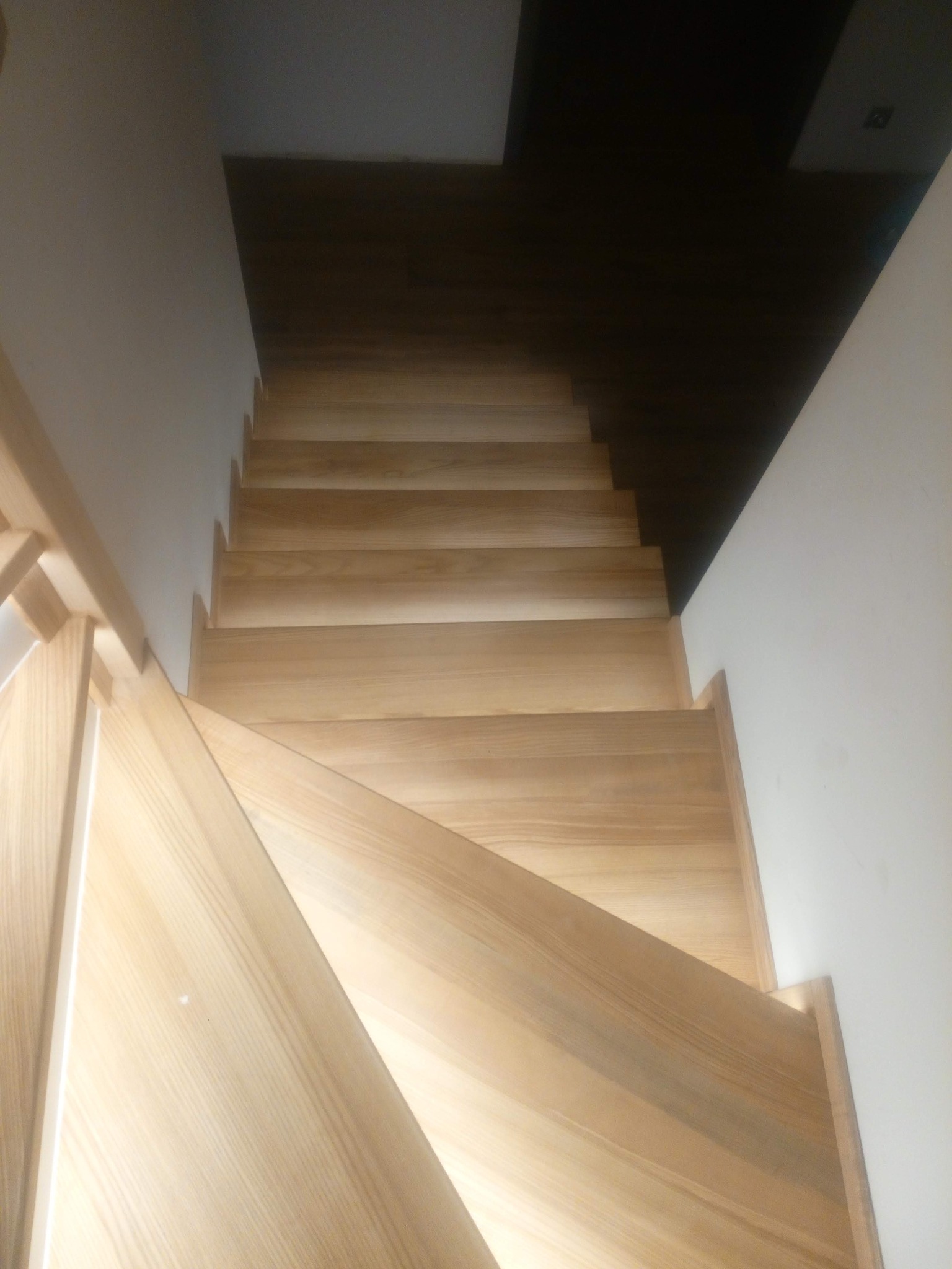 Jasne, drewniane schody z widoczną strukturą drewna, prowadzące w górę, z białymi ścianami po bokach i ciemnym sufitem w tle. Ujęcie z góry, podkreślające naturalny materiał.