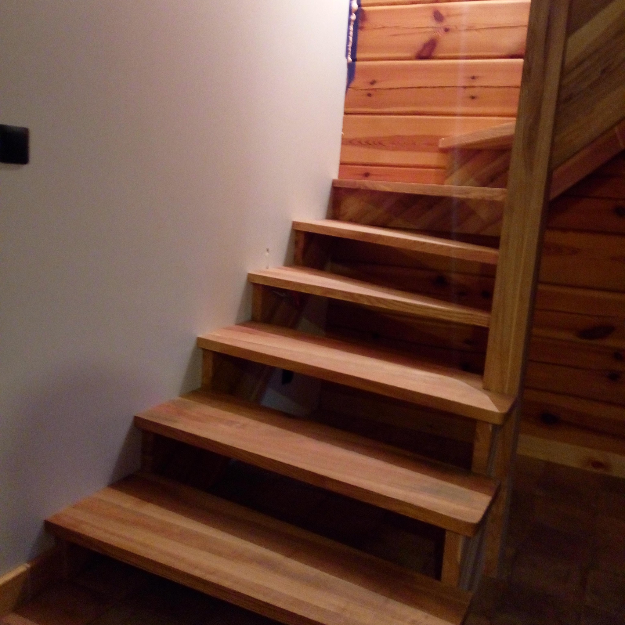 Drewniane schody z jasnego drewna, prowadzące w górę wzdłuż białej ściany i drewnianej boazerii. Widoczne detale konstrukcyjne i naturalne usłojenie drewna.