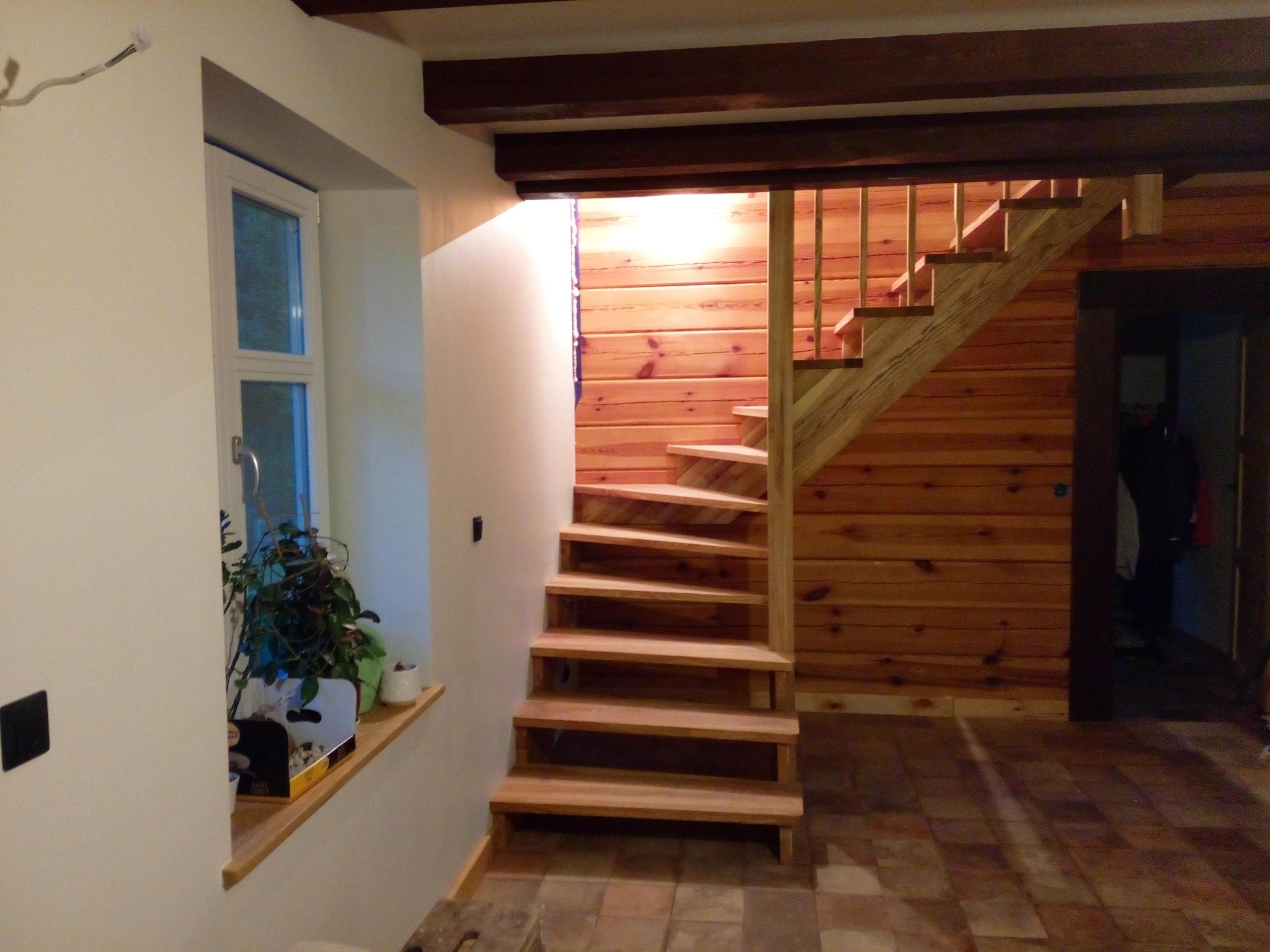 Drewniane schody spiralne z jasnego drewna, prowadzące na górę, z drewnianą balustradą. Ściany wykończone boazerią. Obok okno z roślinami.