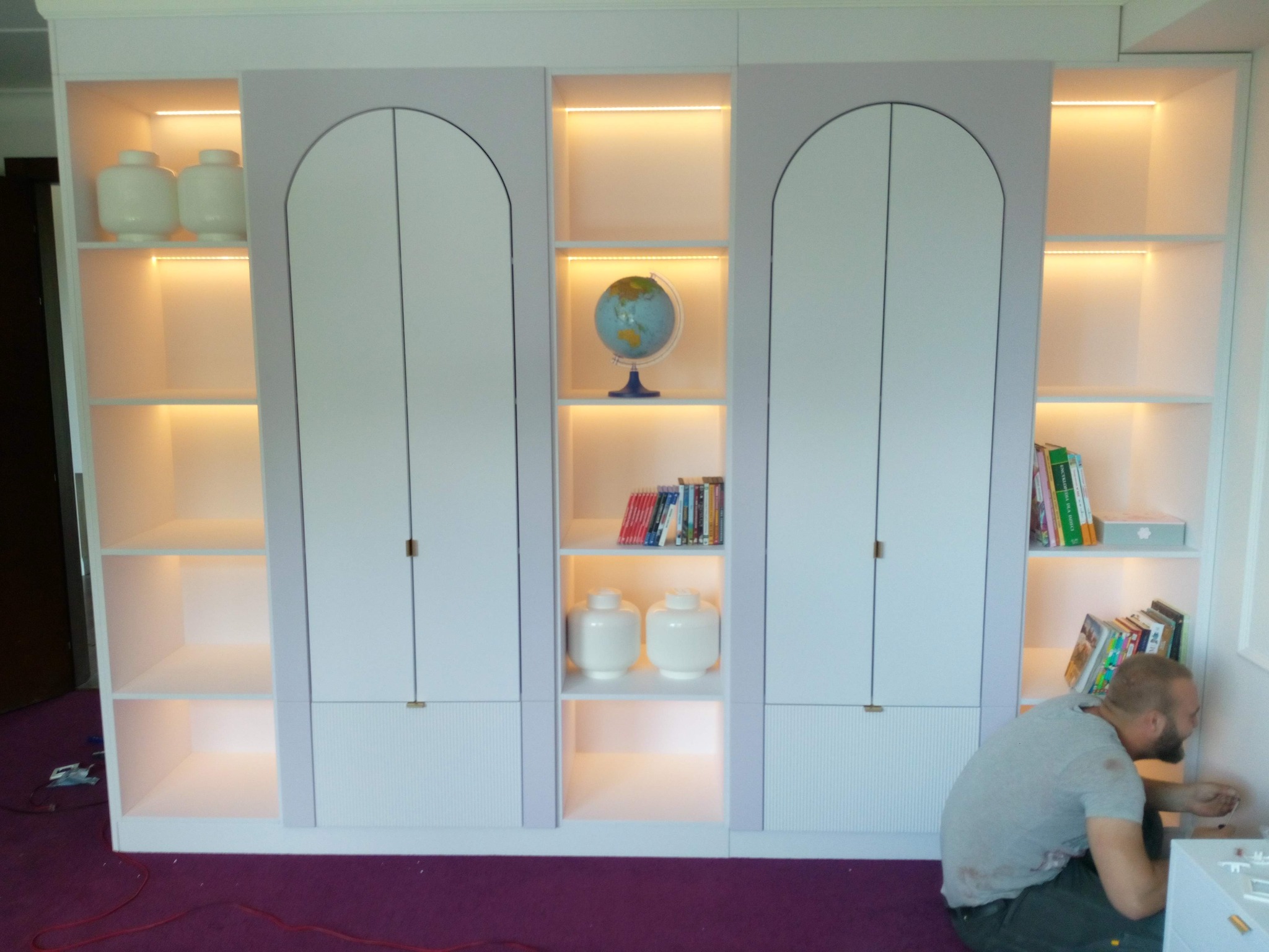 Biała szafa wnękowa z oświetleniem LED i zaokrąglonymi drzwiami, uzupełniona dekoracjami. Montażysta w trakcie pracy, widoczny na pierwszym planie.