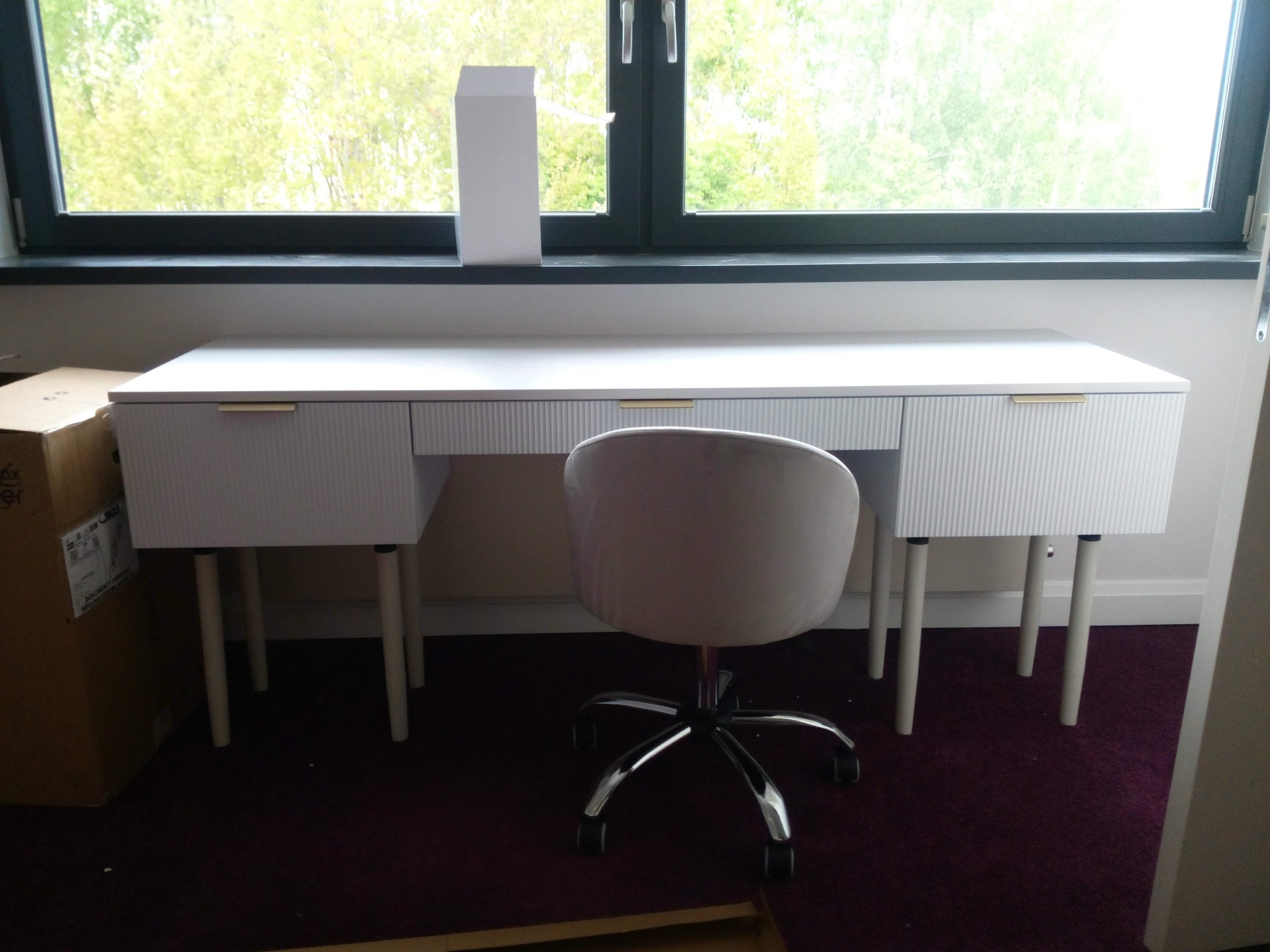 Białe biurko z szufladami i krzesłem na kółkach, ustawione przy oknie. Widoczny karton obok biurka. Minimalistyczny design, jasne wnętrze.