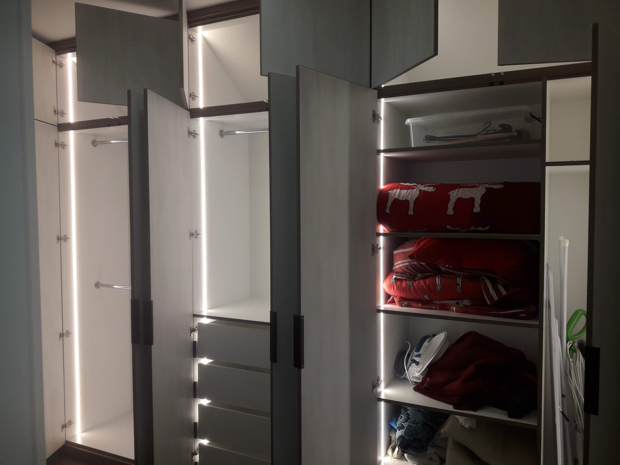Otwarta szafa z szarymi frontami, oświetleniem LED wewnątrz, z widocznymi półkami, szufladami i drążkami na ubrania. Wnętrze szafy wypełnione ubraniami i akcesoriami.