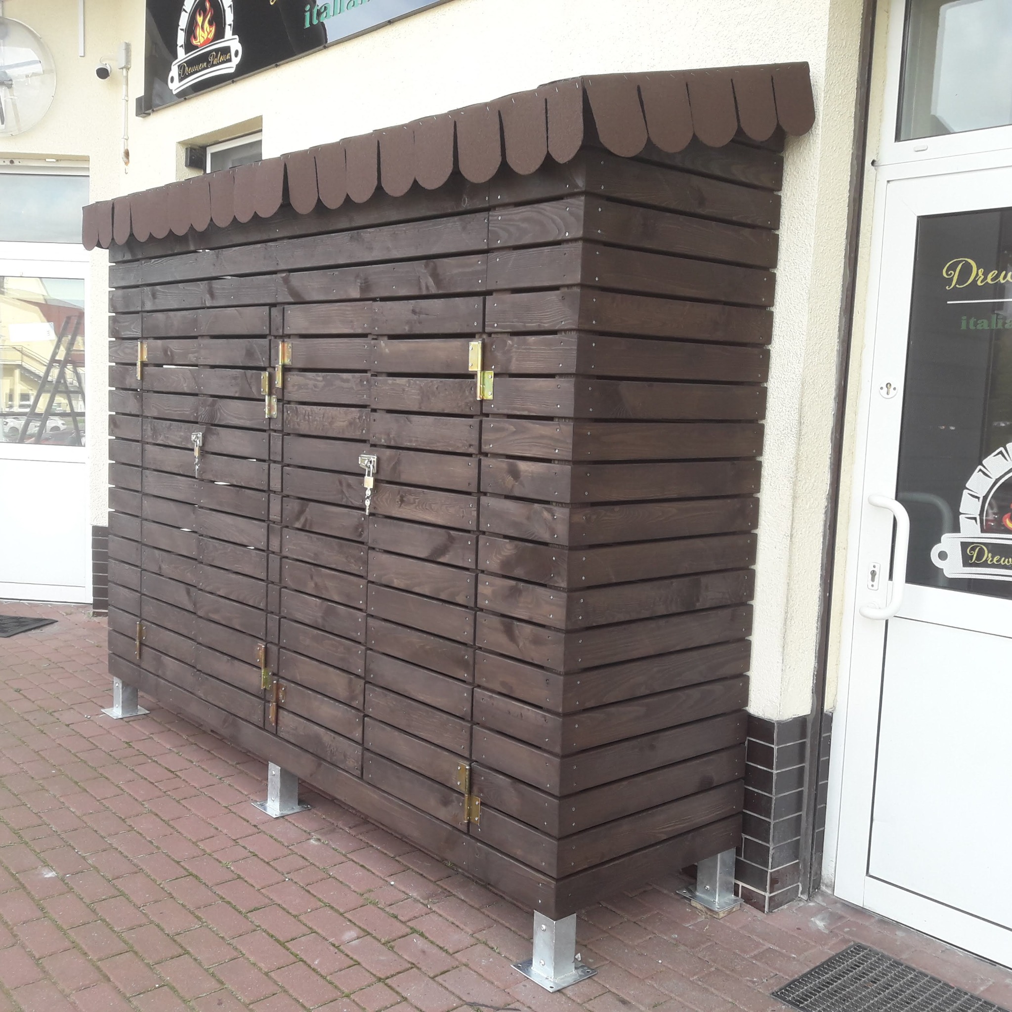 Drewniana zabudowa na zewnątrz budynku, z brązowych desek, zamykana na klucz, z daszkiem. Widoczne metalowe nóżki i ceglana nawierzchnia.