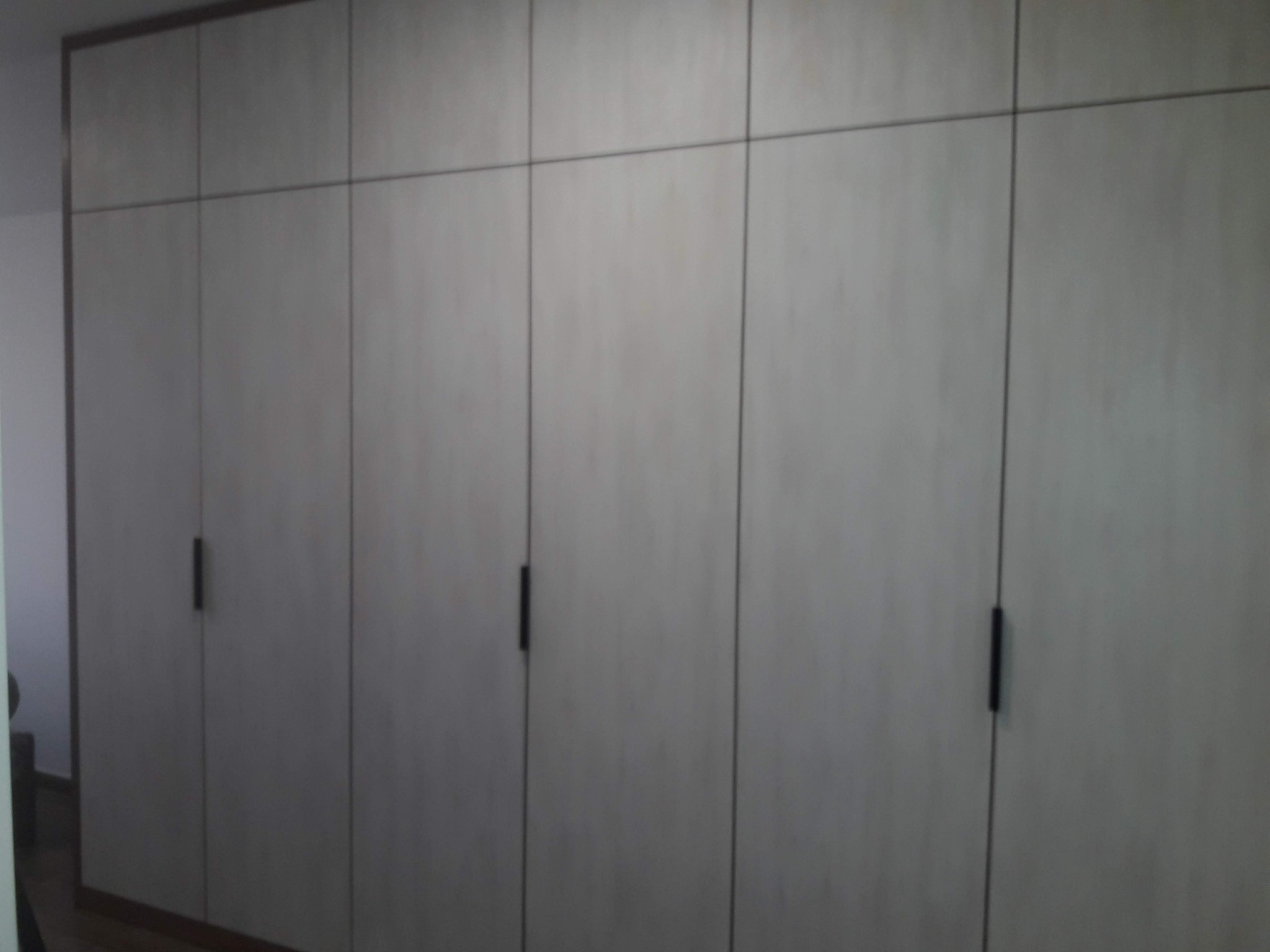 Jasna szafa wnękowa z ciemnymi, minimalistycznymi uchwytami, zabudowana od podłogi do sufitu, z widocznymi podziałami na segmenty.