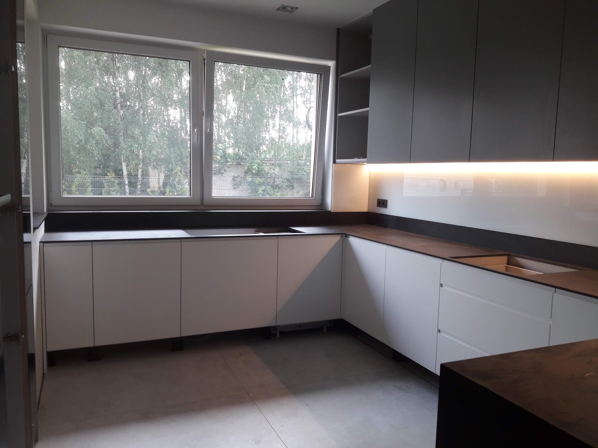 Nowoczesna kuchnia z białymi szafkami dolnymi i szarymi górnymi, podświetleniem LED i widokiem na zieleń za oknem. Minimalistyczny design, ciemny blat.