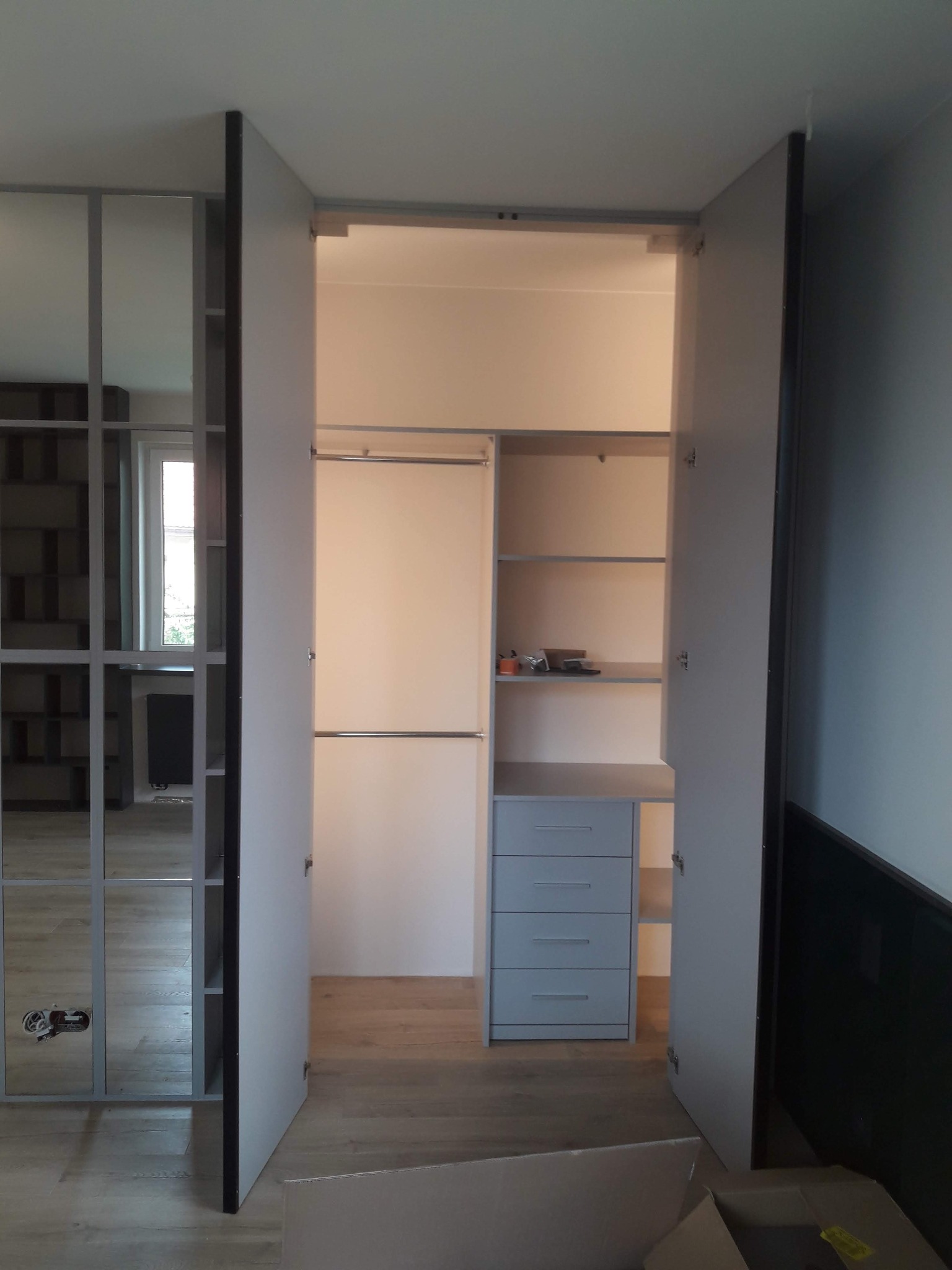 Otwarta szafa wnękowa z szufladami i półkami, oświetlona od wewnątrz, z lustrzanymi drzwiami po lewej stronie. Widoczna podłoga z paneli i fragment ściany.