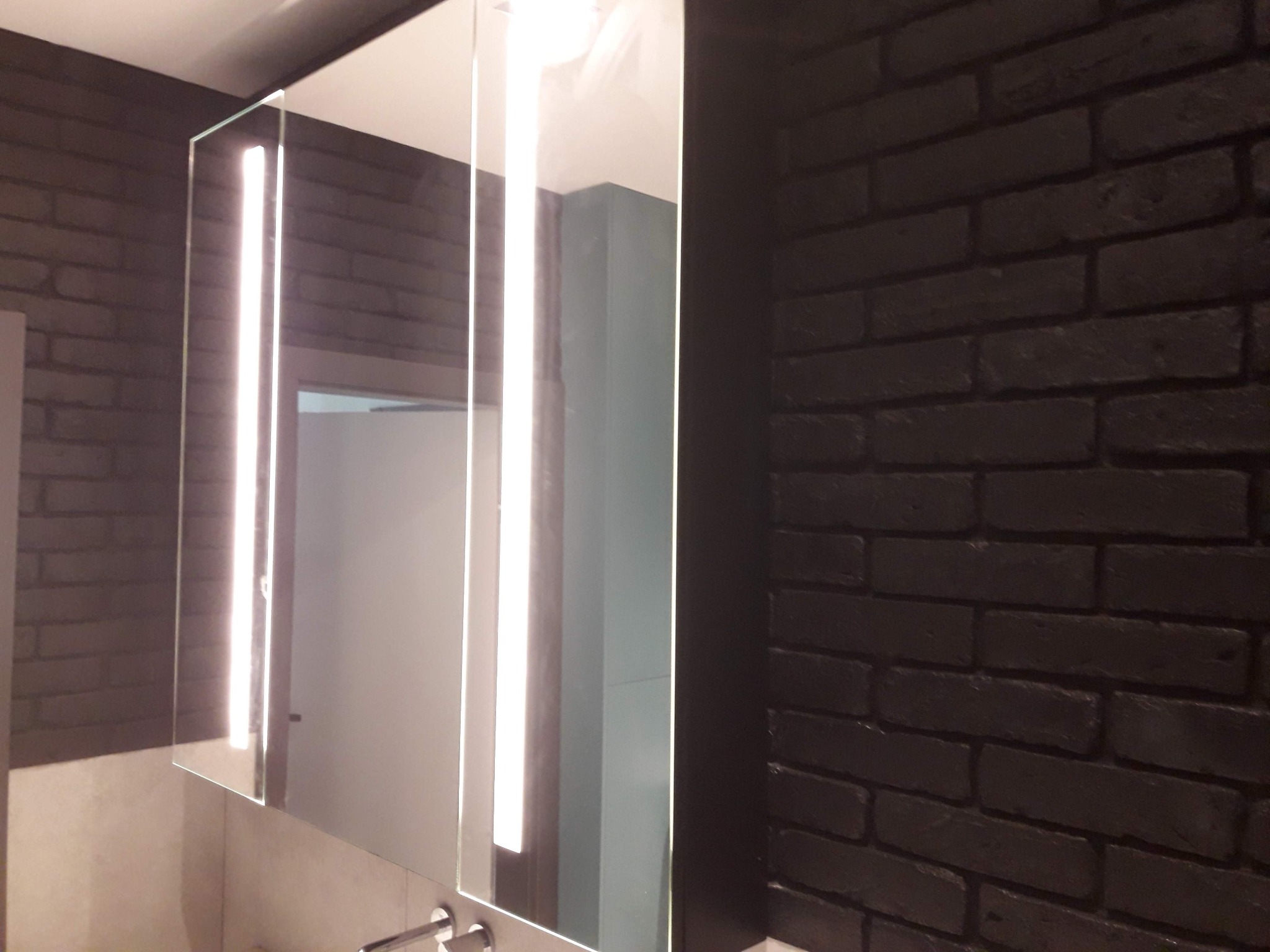 Nowoczesna szafka łazienkowa z lustrem i oświetleniem LED na tle ściany z ciemnej cegły. Minimalistyczny design, eleganckie wykończenie.