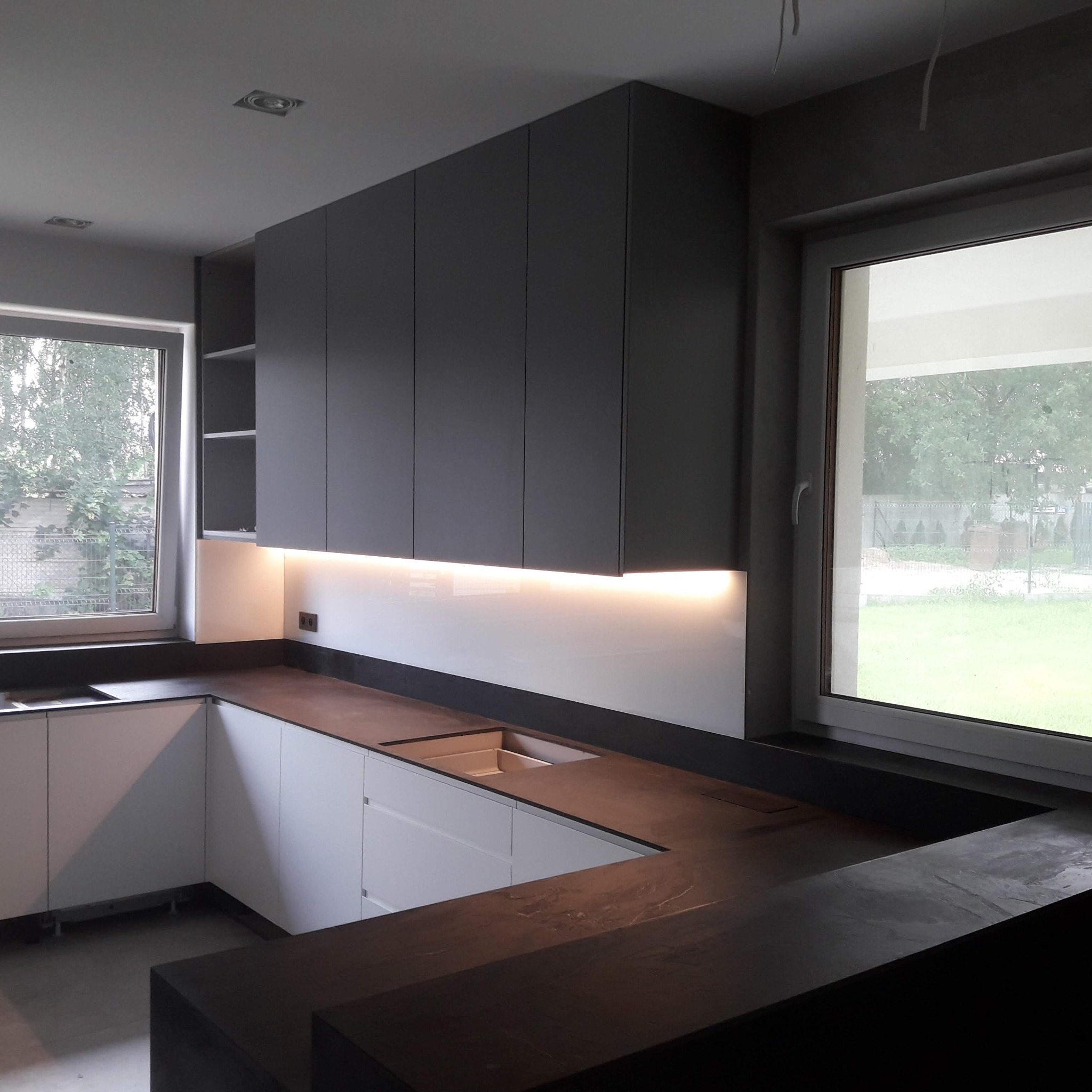 Nowoczesna kuchnia w kształcie litery L z szarymi szafkami wiszącymi i białymi dolnymi, oświetlona listwą LED pod szafkami, z ciemnym blatem.