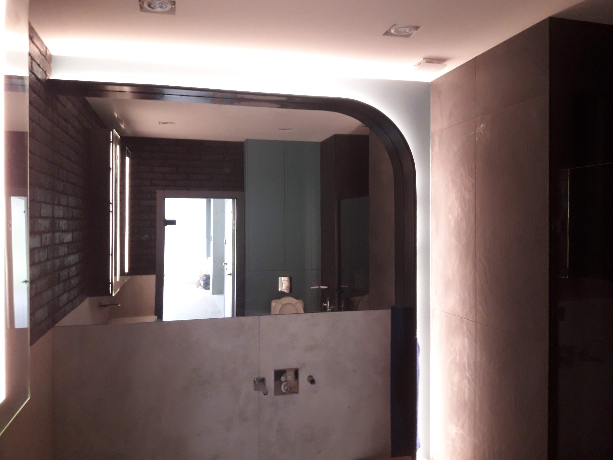 Nowoczesna łazienka z dużym lustrem w czarnej ramie z podświetleniem LED, widok na umywalkę i fragment ściany z cegły. Minimalistyczny design, surowe wykończenie.