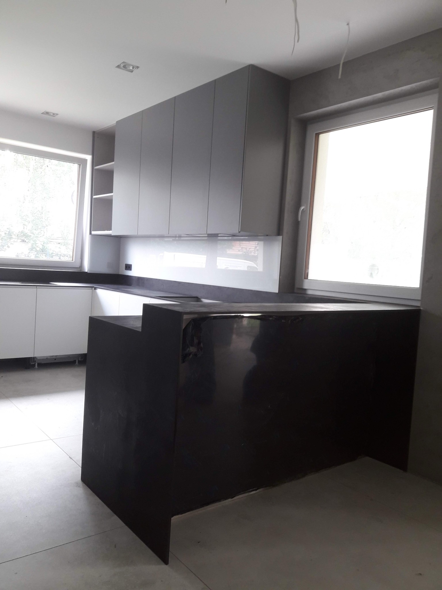 Nowoczesna kuchnia z szarymi szafkami i czarnym blatem, widok na wyspę kuchenną. Minimalistyczny design, jasne wnętrze z oknami.