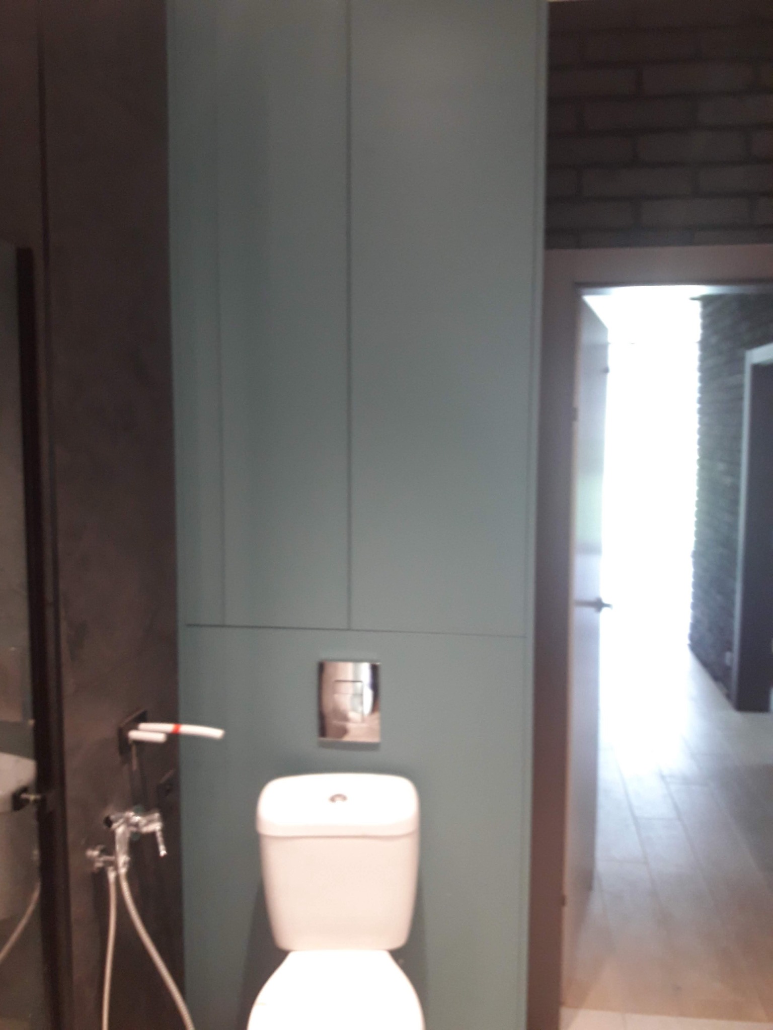 Błękitna zabudowa toalety z szafkami, widoczny sedes i bidetka. W tle ceglana ściana i otwarte drzwi do jasnego pomieszczenia z drewnianą podłogą.