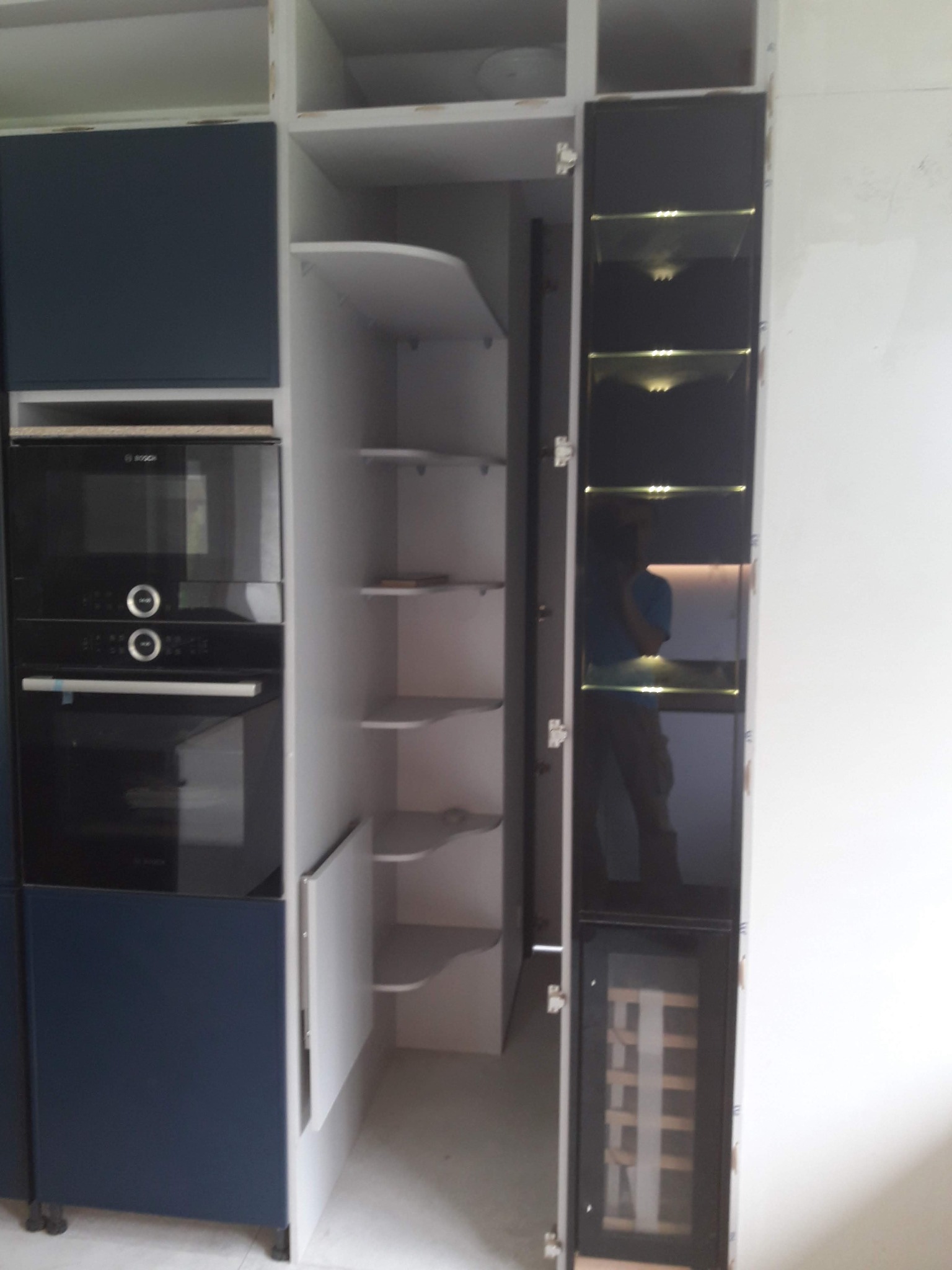 Nowoczesna zabudowa kuchenna z piekarnikiem, szafkami w kolorze granatowym i białym, oraz otwartą szafką z półkami i szafką na wino z czarnymi, szklanymi drzwiami.