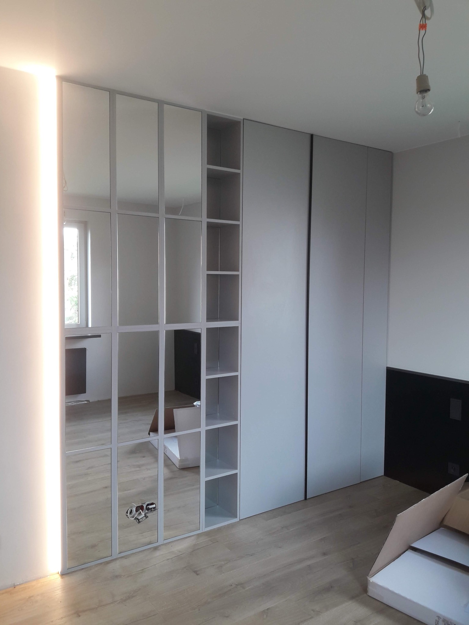 Nowoczesna szafa wnękowa z lustrzanymi drzwiami i otwartymi półkami, oświetlenie LED po lewej stronie, widoczne narzędzia i kartony na podłodze.