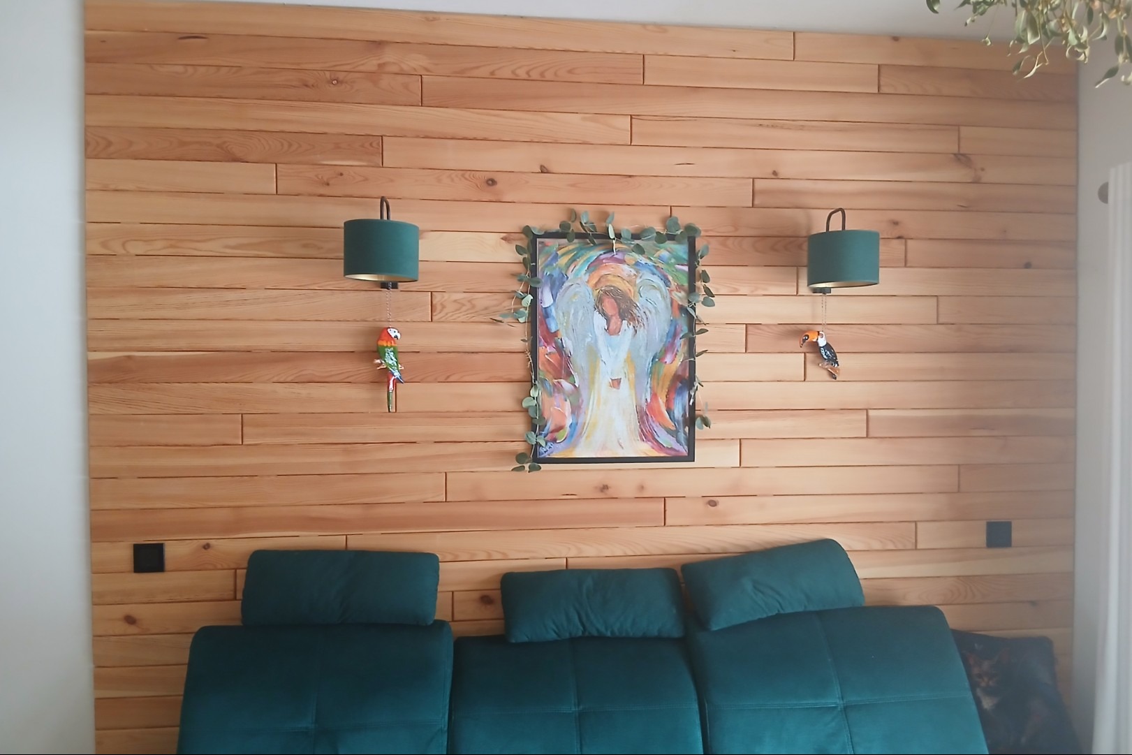 Drewniana ściana z poziomych desek, ozdobiona obrazem w ramie i lampkami z motywem papug. Pod ścianą zielona sofa.