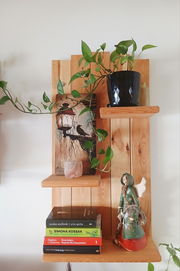 Drewniana półka z desek z trzema poziomami, na których stoją książki, roślina doniczkowa, figurka anioła i obrazek z motywem latarni i kruka. Naturalne drewno, jasne tło.