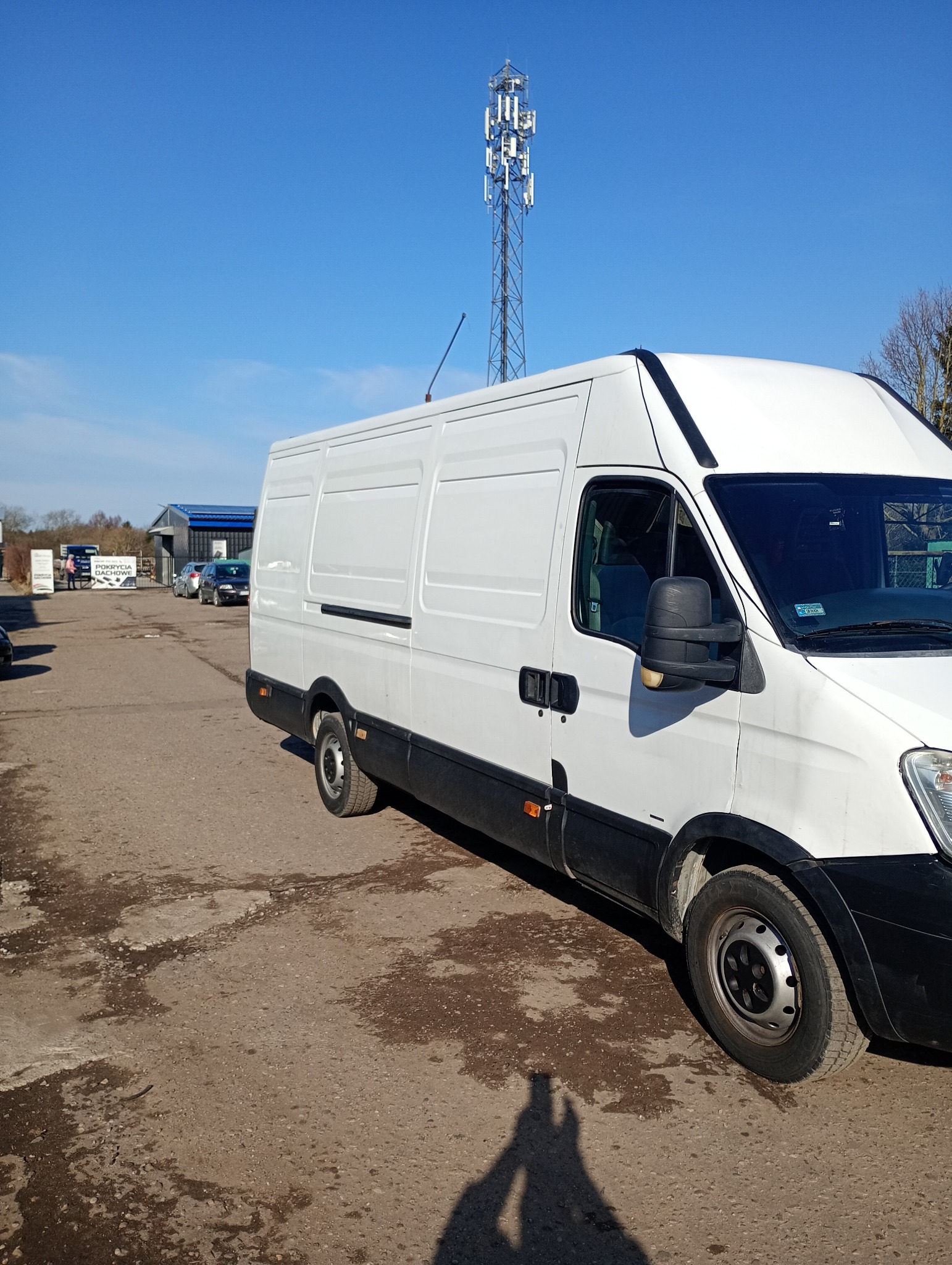 Biały bus dostawczy na placu, z wieżą telekomunikacyjną w tle pod błękitnym niebem. Widoczny cień osoby robiącej zdjęcie.