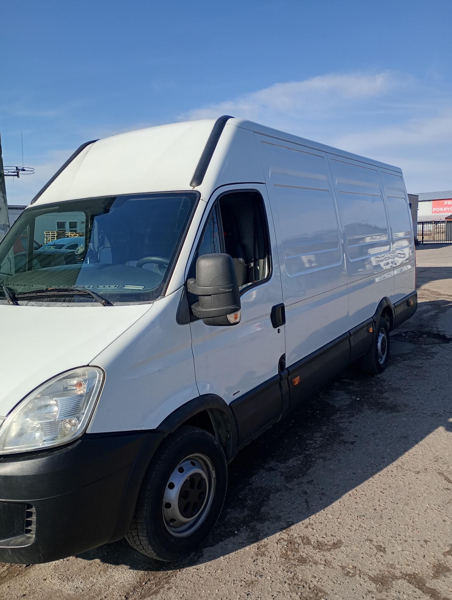 Biały bus dostawczy marki Iveco na betonowym placu, widok z boku, błękitne niebo w tle. Widoczny fragment budynku z reklamą.