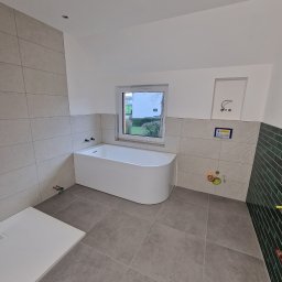 Tomek Zaba - Nowoczesna łazienka z wanną, szarą płytką podłogową i częściowo wykończoną ścianą zieloną glazurą. Widoczne przygotowane instalacje wodne i odpływ liniowy.