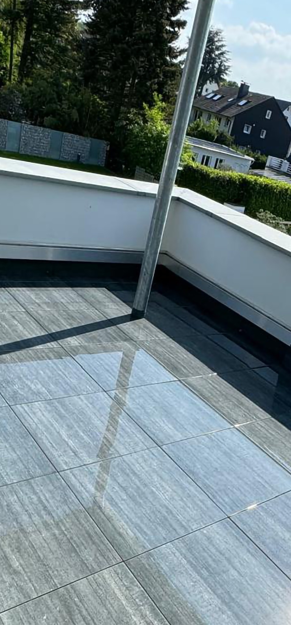 Błyszcząca, szara, ceramiczna podłoga balkonu z widocznym słupem i fragmentem białej balustrady. W tle zieleń i zabudowa jednorodzinna.