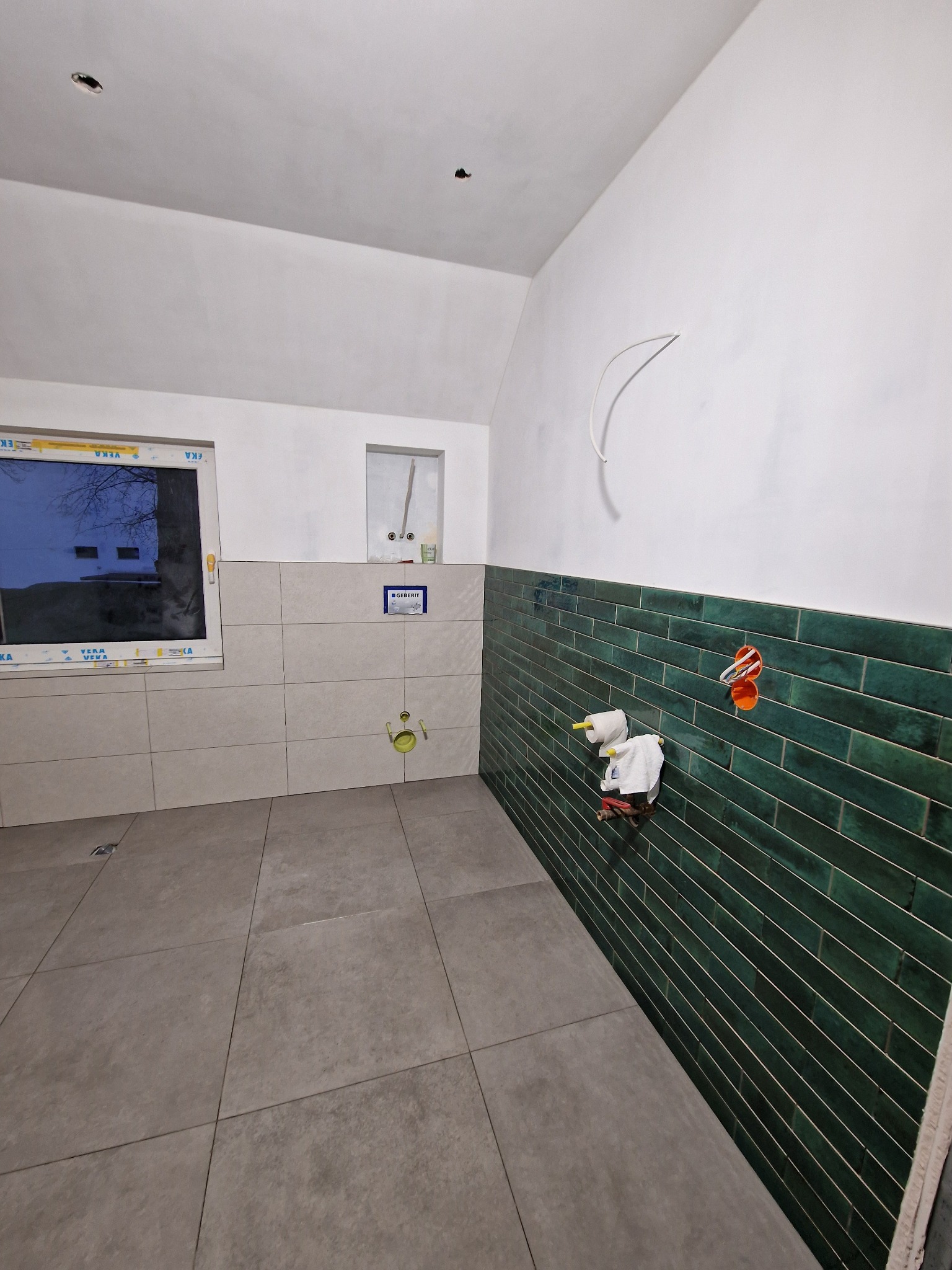 Wnętrze łazienki w trakcie wykańczania: ułożone szare płytki podłogowe i zielone płytki ścienne do połowy wysokości. Widoczne instalacje i okno z widokiem na zewnątrz.