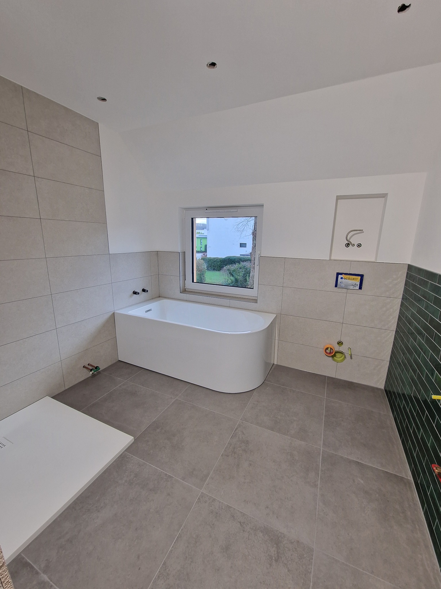 Nowoczesna łazienka z wanną, szarą płytką podłogową i częściowo wykończoną ścianą zieloną glazurą. Widoczne przygotowane instalacje wodne i odpływ liniowy.