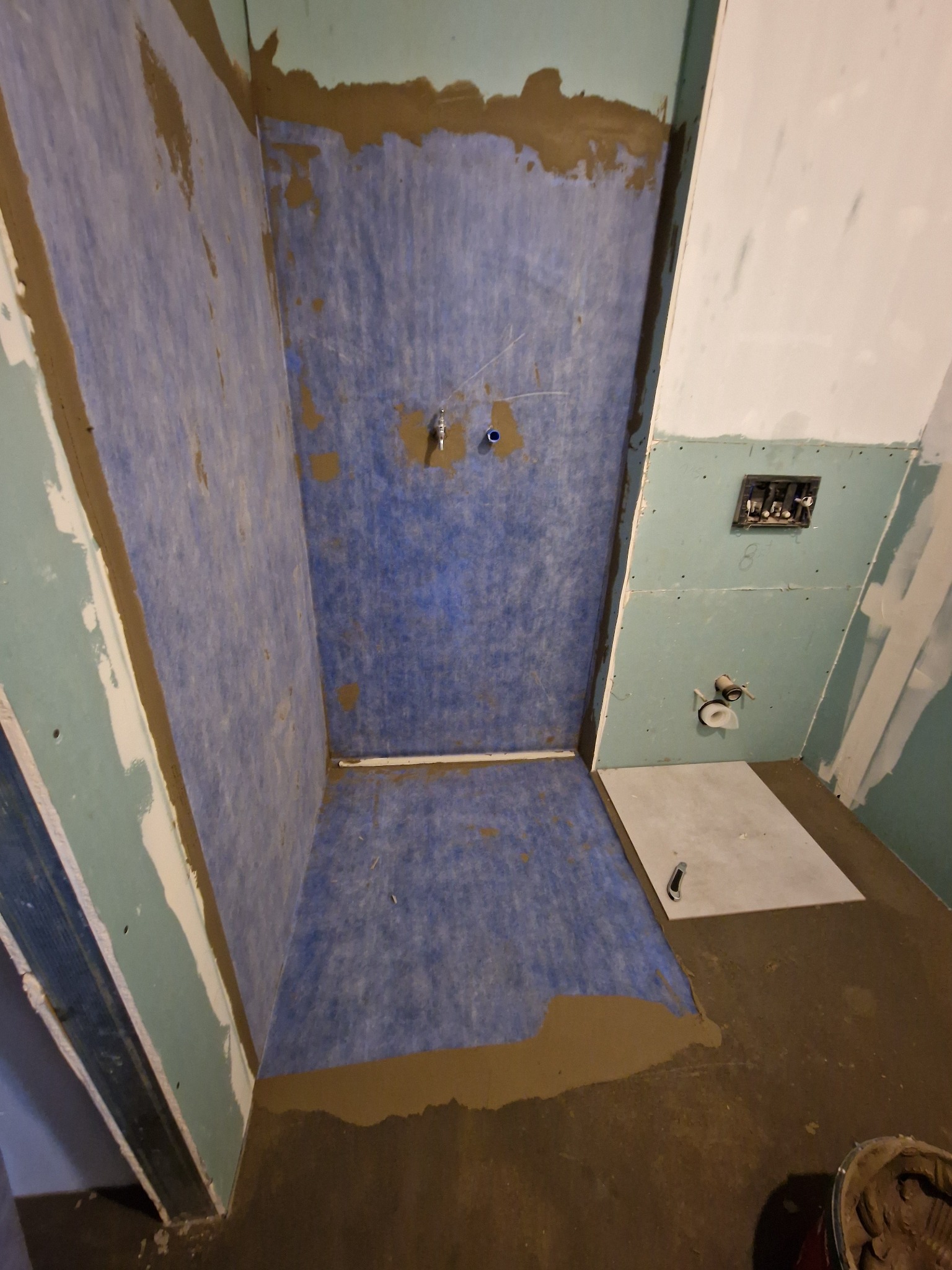 Przygotowanie do hydroizolacji w kabinie prysznicowej: niebieska membrana w płynie na ścianach i podłodze, widoczne instalacje wodne i elektryczne, surowy stan wykończenia.