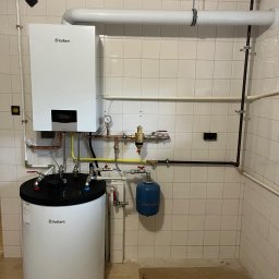 Aquapro Krzysztof Himik - Biały piec gazowy Vaillant z zasobnikiem wody, rurami miedzianymi i czarnymi, na tle wykafelkowanej ściany. Widoczne zawory i manometr.