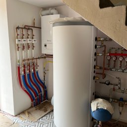 Aquapro Krzysztof Himik - Biały zasobnik ciepłej wody użytkowej Vaillant z podłączeniami, obok piec gazowy, instalacja z rur miedzianych i liczniki. Widok w kotłowni pod schodami.