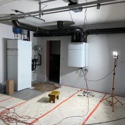 Aquapro Krzysztof Himik - Biały piec gazowy Vaillant zamontowany na ścianie w kotłowni, widoczne rury, instalacja w trakcie montażu, pomarańczowe linie na podłodze, lampa oświetleniowa.