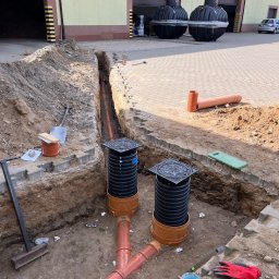Aquapro Krzysztof Himik - Montaż instalacji kanalizacyjnej z rur PCV w wykopie ziemnym, widoczne studzienki rewizyjne z pokrywami. W tle budynek z dwoma zbiornikami na szambo.