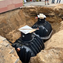 Aquapro Krzysztof Himik - Dwa czarne zbiorniki na szambo umieszczone w wykopie ziemnym, zabezpieczone płytami. Widoczny fragment budynku i osoby obserwujące prace.