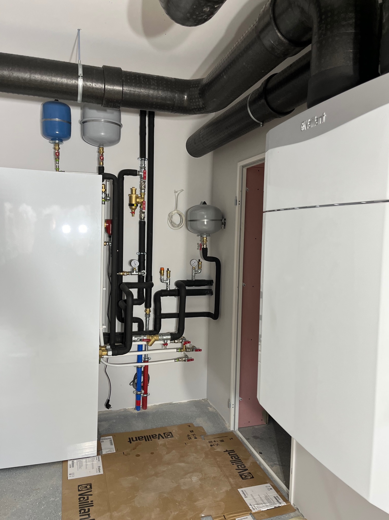 Nowoczesna kotłownia z piecem gazowym Vaillant, rurami instalacji grzewczej i zbiornikami wyrównawczymi. Widoczne elementy instalacji w kolorach czarnym, czerwonym i niebieskim.