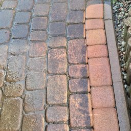 Umyję Umebluję Kamil Klonowski - Częściowo umyta kostka brukowa w odcieniach brązu, widoczna różnica w czystości, mokra powierzchnia, obrzeże z kostki i kamieni, trawa w tle.