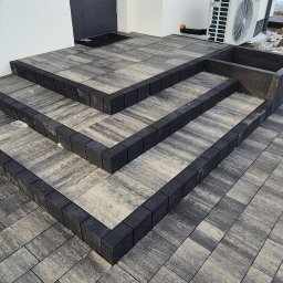 Umyję Umebluję Kamil Klonowski - Schody i chodnik z kostki brukowej w odcieniach szarości, z czarnym obramowaniem, prowadzące do białego budynku. Widoczny fragment klimatyzatora i resztki śniegu.