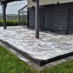 Kostka brukowa na tarasie w trakcie czyszczenia myjką ciśnieniową. Widoczne ślady piany i czyszczona powierzchnia. W tle dom z ciemną elewacją i zadbany trawnik.