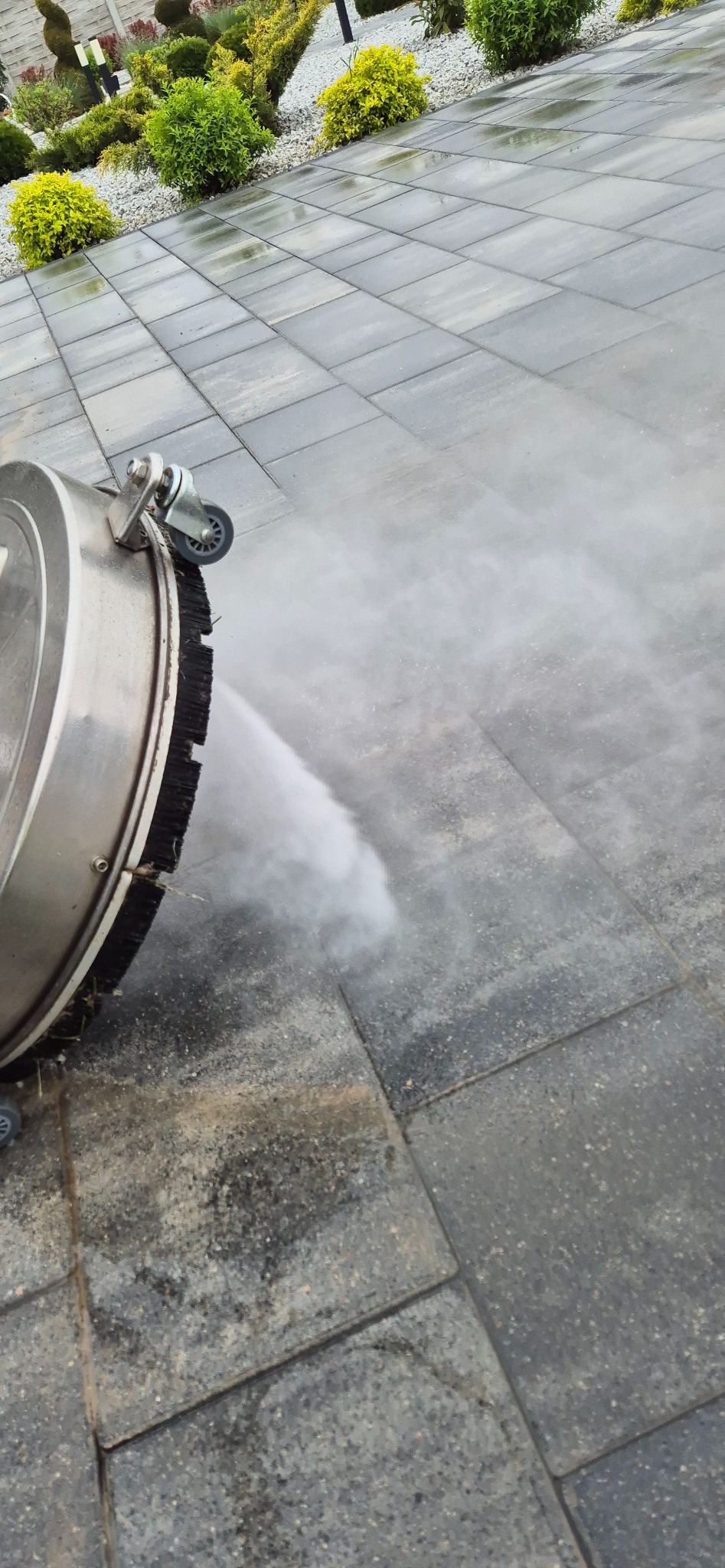 Szczotka rotacyjna pod ciśnieniem czyści szare kostki brukowe, z boku widoczne rośliny ozdobne. Woda pod ciśnieniem usuwa brud z powierzchni kostki.