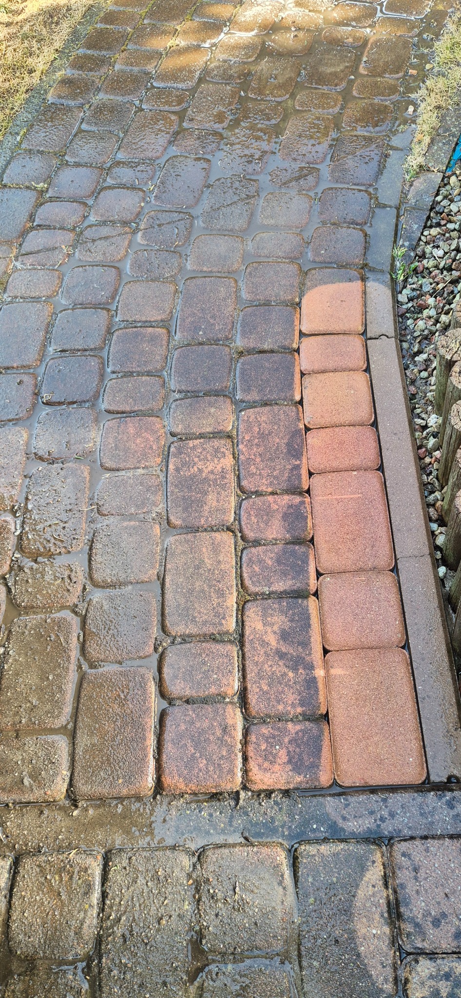 Częściowo umyta kostka brukowa w odcieniach brązu, widoczna różnica w czystości, mokra powierzchnia, obrzeże z kostki i kamieni, trawa w tle.
