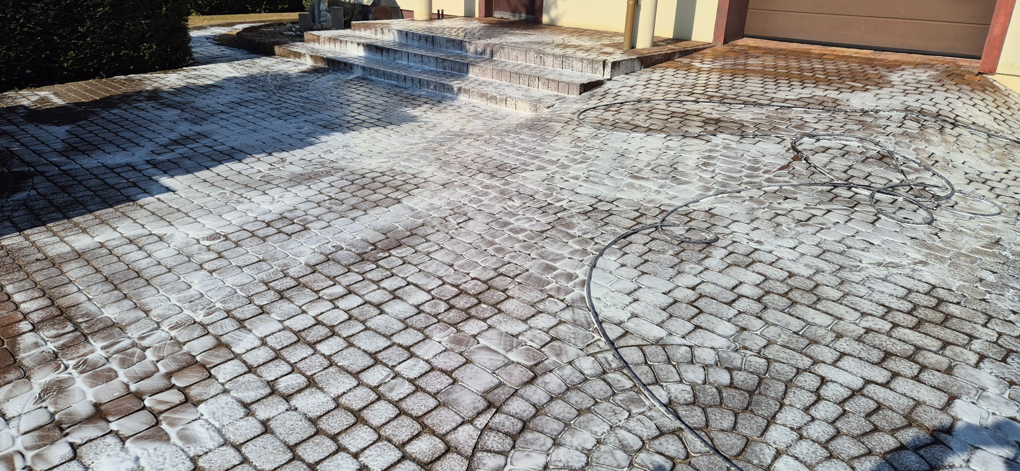 Czyszczona kostka brukowa przed domem z użyciem piany aktywnej. Widoczne schody wejściowe i częściowo zwinięty wąż ciśnieniowy. Jasne światło słoneczne.